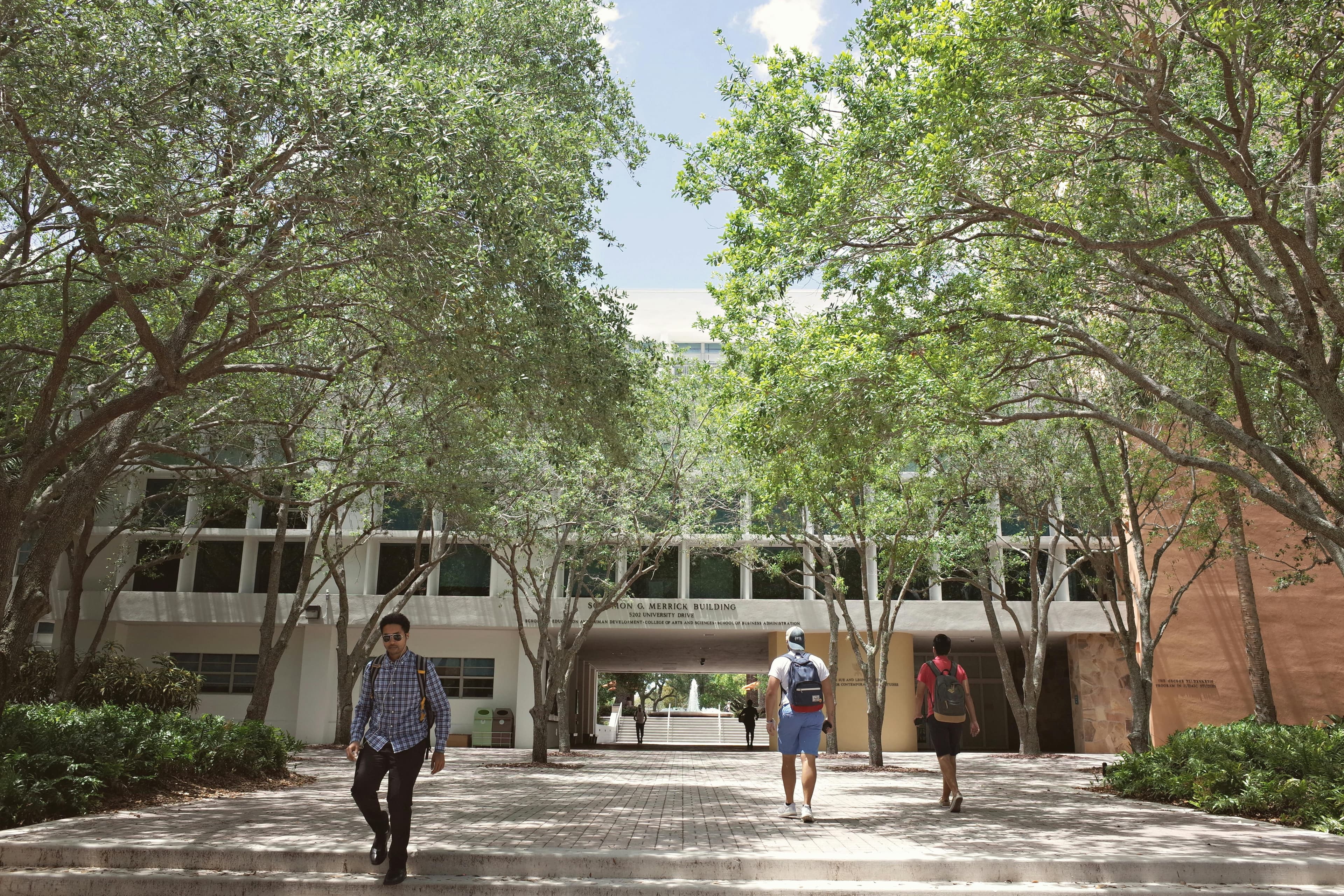 Students walking through a tree-shaded campus walkway between modern academic buildings.