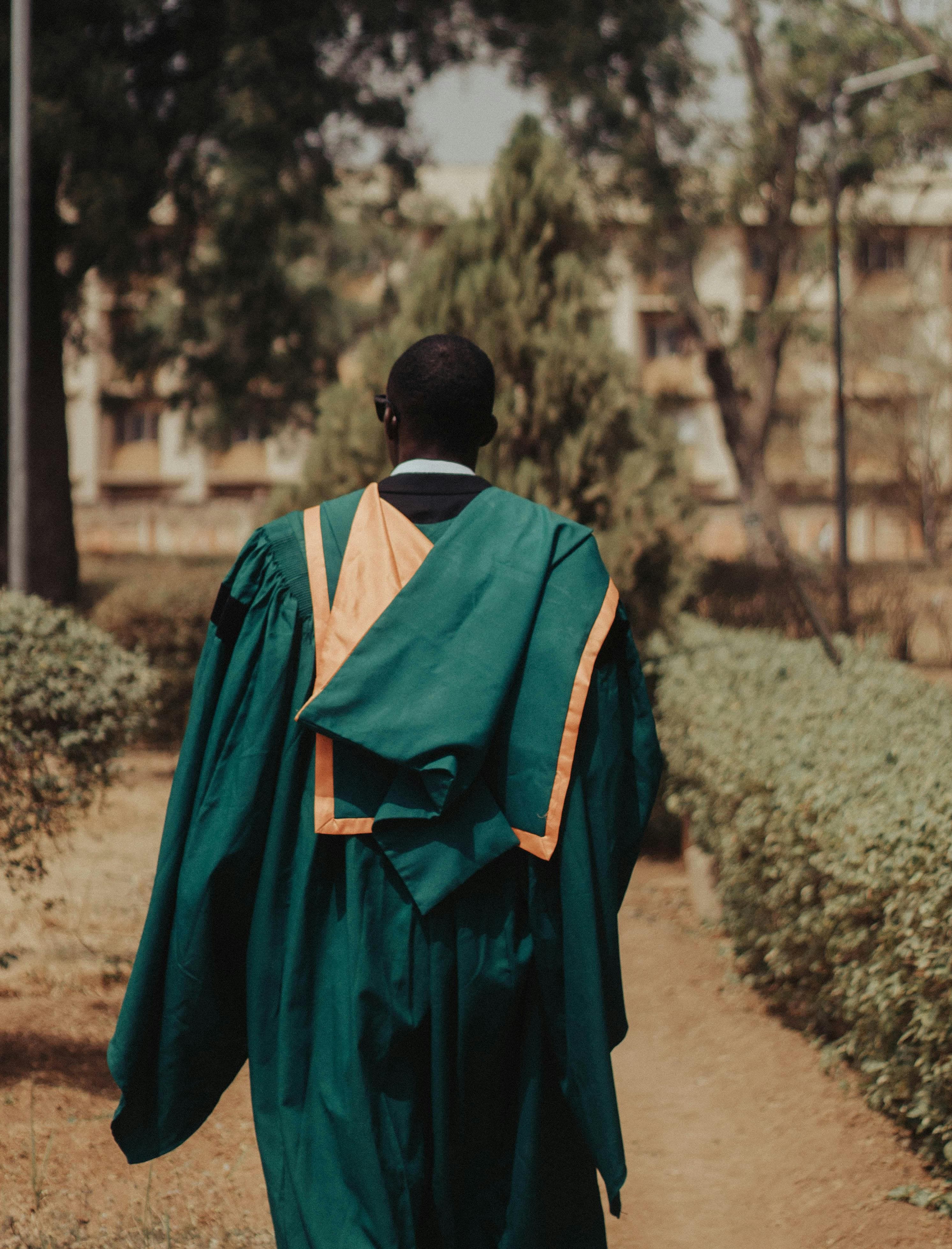 Graduate in green and gold academic regalia walking on a campus path, photographed from behind.
