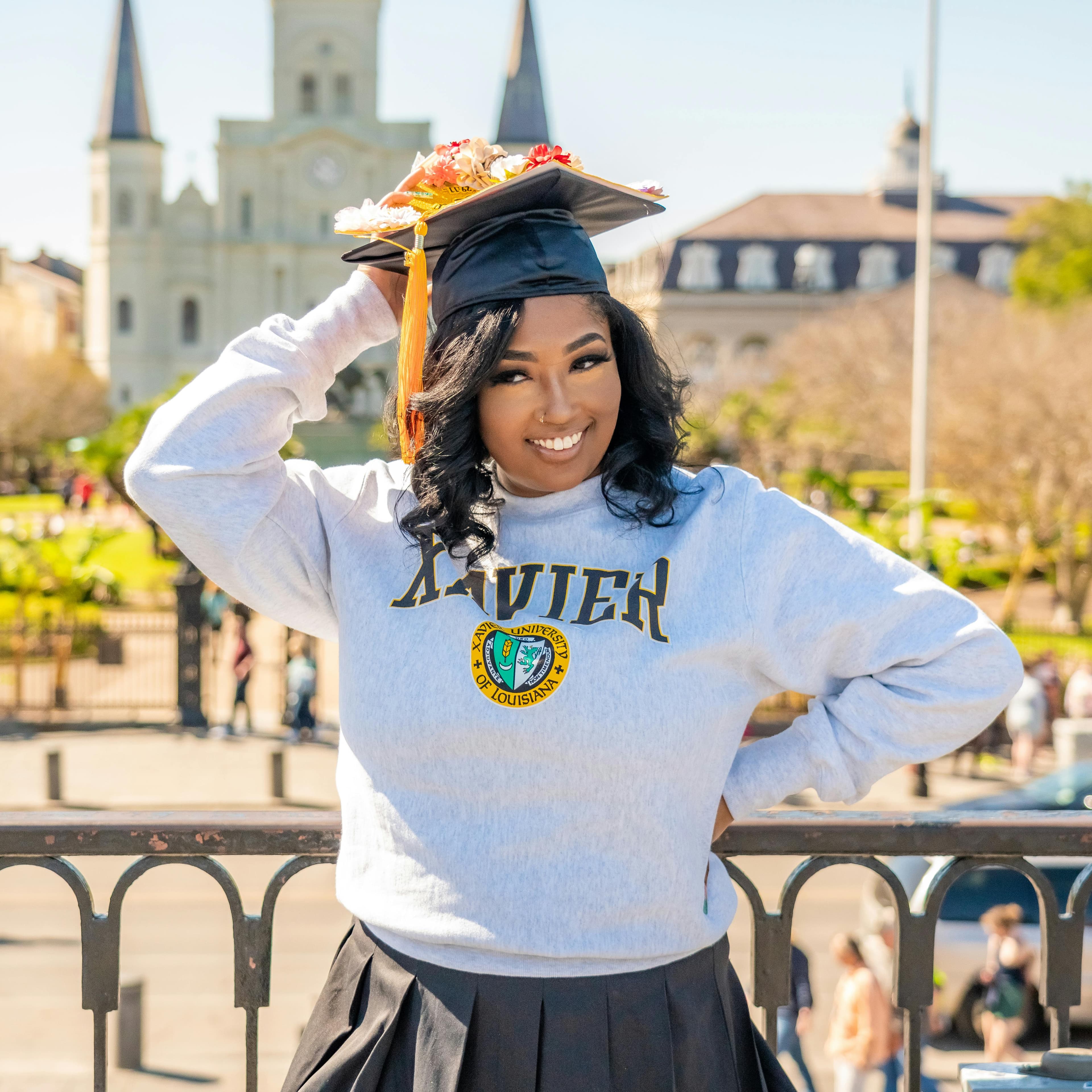 Smiling graduate in academic regalia with floral cap decoration, photographed in front of a historic campus building.